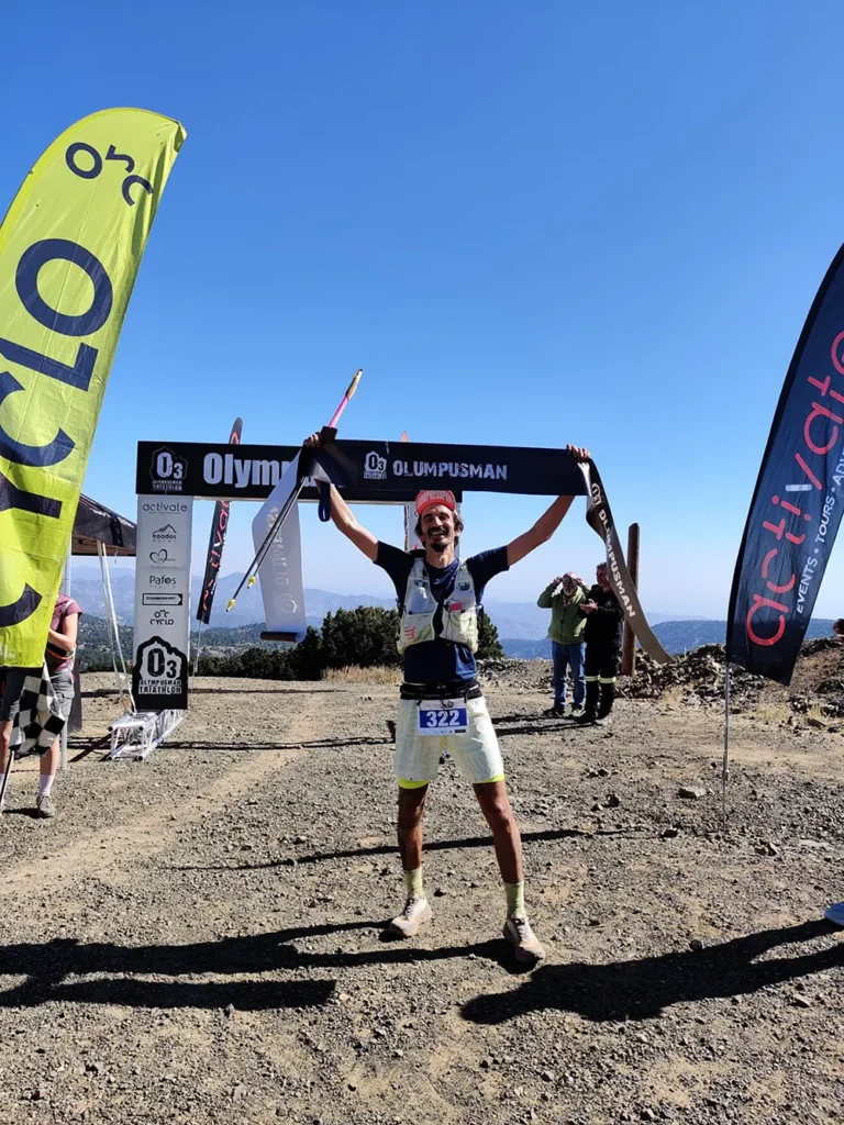 Endurance athlete Viktor Olov Leonidou holding finish line banner at OlympusMan race, with Raw + Tonic branding visible in background.